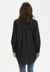 Sort langærmet skjorte lavet i glat stof, med rundet kant, rygplissé og manchetter på ærmerne. Enkel og strømlinet design.