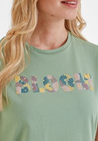 Light green cotton t-shirt featuring the word "BLOOM" in colorful floral patterns. Round neck and soft texture. Close-up view of the print.