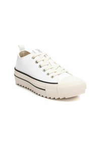 Zapatilla de lona blanca con una puntera de goma, suela gruesa y texturizada, y detalles de franjas negras alrededor de la mediasuela. Los cordones cuentan con ojetes de metal.