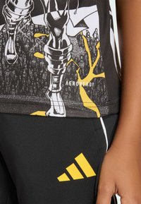 Sort atletisk top med grafisk print og gule accenter, parret med sorte bukser med en fremtrædende gul Adidas-logo. Materialet ser ud til at være strækbart.