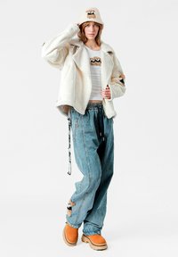 Chaqueta blanca con forro de borrego, camiseta blanca con gráfico de oso, pantalones vaqueros azules de pierna ancha con aplique de oso, zapatos naranjas sin cordones y sombrero tipo cubo de punto.