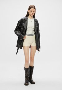 Sort læderjakke med struktureret finish, hvid tætsiddende skjorte, beige shorts med logo-betræk, og knæhøje sorte støvler med spænder.