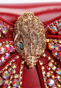Gouden handvormige broche versierd met bruine kristallen, bevestigd aan een rode stof versierd met multikleurige strass en kraembroderie.