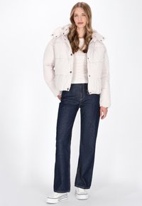 Hellbeige Daunenjacke mit Kapuze, mit einem gesteppten Design und silbernen Druckknöpfen, kombiniert mit dunkelblauen geraden Jeans und weißen Sneakers.