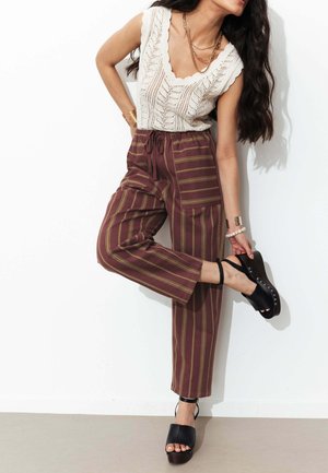 Femme ajustant des sandales compensées noires, portant un haut sans manches en maille blanche et un pantalon marron rayé, debout contre un mur blanc.