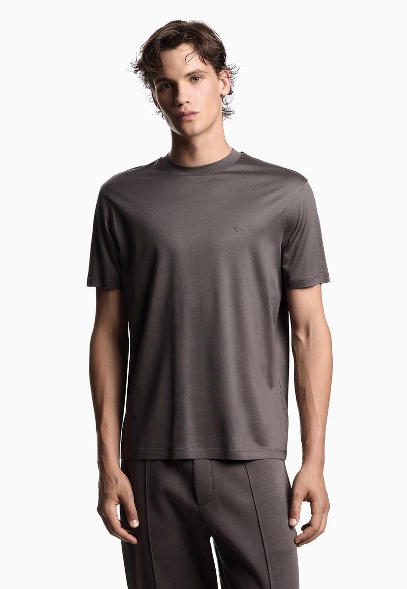 Jonge man in een effen donkergrijs T-shirt met korte mouwen en bijpassende broek, staande tegen een witte achtergrond.