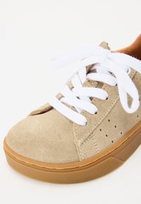 Beige mockaläder sneakers med vita snören, vadderad tunga och gummisula, med perforerade detaljer längs sidan för ventilation.