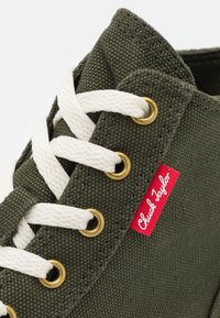 Primo piano di una sneaker in tela verde oliva con lacci bianchi infilati attraverso occhielli di ottone e un'etichetta in tessuto rossa "Chuck Taylor".