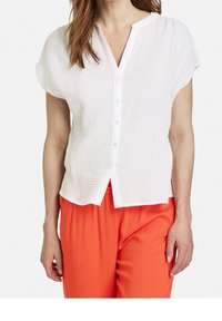 Femme portant un chemisier blanc à manches courtes et texturé, associé à un pantalon ample orange vif.