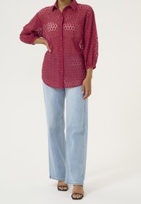 Červená krajková košile na knoflíky s vzorem včelí plástve, spárovaná se světlými modrými širokými džínami a černými sandály na otevřenou špičku.