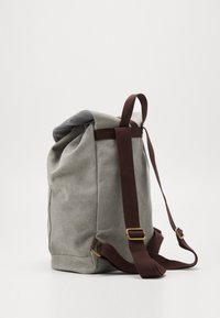 Sac à dos en toile grise avec des bretelles réglables en brun foncé et une poignée de boucle, se tenant debout sur un fond blanc.