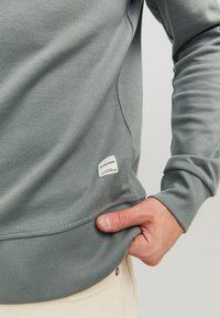 Grauer Pullover mit geripptem Saum und Bündchen, ausgestattet mit einem weißen Etikett auf der unteren Seite. Der Stoff wirkt glatt und hat einen leichten Glanz.