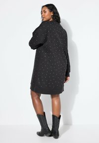 Schwarzes, übergroßes Sweatshirtkleid mit weißen Nietenverzierungen, ausgestattet mit langen Ärmeln und einem gerippten Saum. Kombiniert mit kniehohen schwarzen Cowboystiefeln.