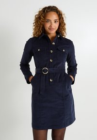 Femme aux cheveux bouclés portant une robe bleu marine à boutons avec des poches poitrine et une ceinture assortie, les mains dans les poches latérales, debout contre un fond blanc.