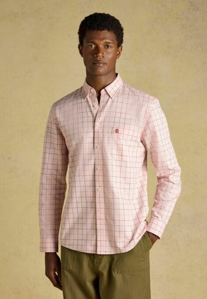 Uomo che indossa una camicia rosa chiaro a maniche lunghe con motivo a griglia rossa e pantaloni verde oliva, in piedi su uno sfondo beige.