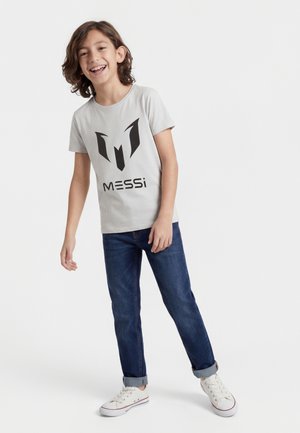 Niño sonriente con cabello de longitud media, vestido con una camiseta gris claro con el logo de Messi, jeans azul oscuro remangados y zapatillas blancas.