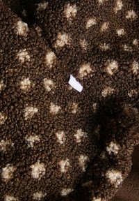 Tessuto di peluche marrone con superficie morbida e strutturata, decorato con motivi floreali color crema e una piccola etichetta nell'angolo.