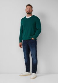 Grüner V-Ausschnitt-Pullover aus weichem Strickstoff, kombiniert mit dunkelblauen Jeans und weißen Turnschuhen. Modell steht vor einem schlichten Hintergrund.