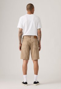 Beige denim shorts met een relaxed fit, zichtbare achterzakken en een bruine leren patch. Gemaakt in combinatie met een effen wit T-shirt en zwarte sneakers.