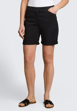 Femme portant un short noir à ourlet roulé, un haut blanc et des sandales plates noires sans bride, debout avec la main droite dans la poche et la main gauche détendue.