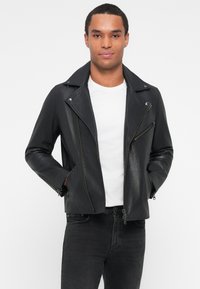 Chaqueta de motocicleta de cuero negro con solapa ancha, cremalleras frontales asimétricas y bolsillos con cremallera. Lleva puesta sobre una camiseta blanca.