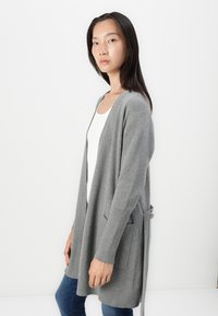 Szary sweter robiony na drutach z otwartym przodem, ściągaczami na rękawach, dwoma przednimi kieszeniami i detalem w postaci paska w talii. Uzupełniony białą bluzką i niebieskimi dżinsami.