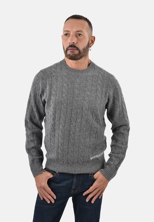 Maglione grigio a maglia con motivo a treccia, polsini e orlo a costine, e testo ricamato in basso. Indossato con jeans blu scuro.