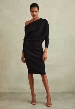 Nő pózol fekete bordázott kötött váll nélküli ruhában és bézs nyitott orrú magassarkúban semleges, egyszínű háttér előtt.