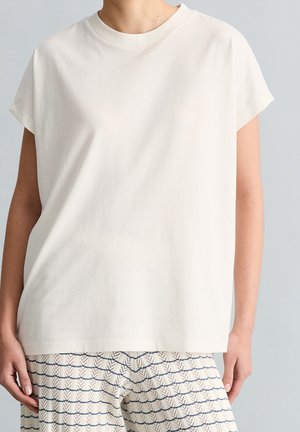 Persoon die een effen wit kortärmelig shirt en losse geruite broek met een blauw-wit patroon draagt tegen een lichte achtergrond.