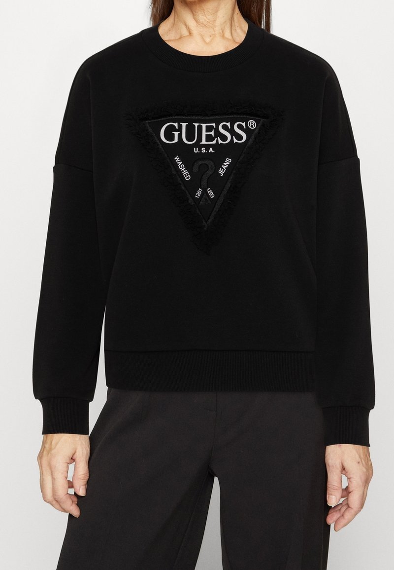 Sudadera negra con un logo triangular de textura negra en relieve, "GUESS U.S.A" y un signo de interrogación. Puños y dobladillo acanalados.