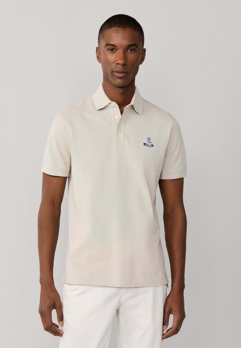 Mann trägt beige kurzärmliges Poloshirt mit kleinem Emblem auf der Brust und weiße Hose, steht vor einfachem hellgrauem Hintergrund.