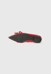 Chaussure à talon haut rouge vue par le dessous, présentant un design élégant et des détails de nœud élaborés près de l'avant, sur un fond blanc.