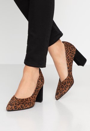 Femme portant des talons hauts à imprimé léopard marron et noir avec talons carrés et pantalon noir sur fond blanc.