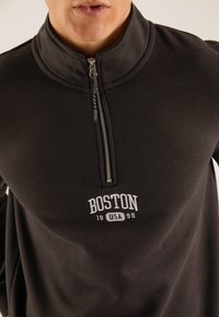 Sudadera negra con cremallera y cuello alto, que presenta "BOSTON 1998" bordado en blanco en el pecho y un detalle de cremallera metálica.