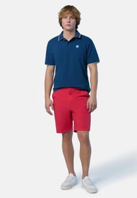 Polo navy con accenti gialli, abbinato a pantaloni corti rossi. Il modello indossa sneakers bianche, mostrando un look estivo informale.