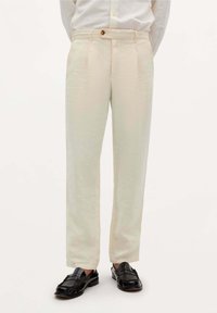 Hellbeige Hose aus strukturiertem Stoff mit Vorderfalten und einem einzigen Knopfverschluss. Dazu schwarze Slipper.
