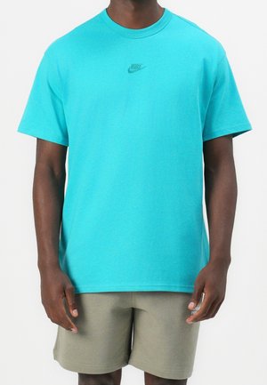 Mand iført en turkis Nike T-shirt og olivengrønne shorts, stående med armene afslappet ned langs siden mod en hvid baggrund.