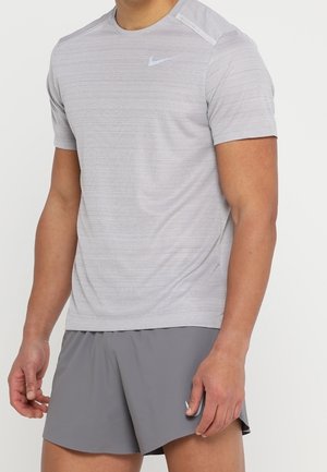 Mężczyzna w jasnoszarej koszulce Nike z krótkim rękawem i szarych sportowych szortach Nike, stoi z rękami swobodnie opuszczonymi wzdłuż ciała na białym tle.