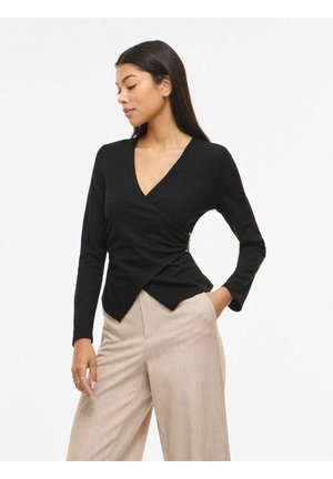 Kobieta o długich ciemnych włosach, ubrana w czarną asymetryczną kopertową bluzkę i beżowe spodnie z szerokimi nogawkami, pozująca z jedną ręką w kieszeni.