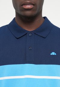 Mörkblå pikétröja med krage, med ljusblå horisontella ränder och en liten blå logotyp på bröstet. Tre-knapps knäppning.