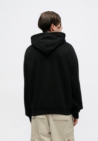 Czarny oversize hoodie z kapturem na sznurki, z miękkiego materiału i ściągaczami przy mankietach, widoczny z tyłu, w połączeniu z jasnobeżowymi spodniami cargo.