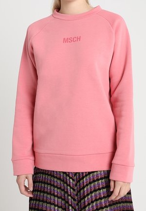 Kvinde iført en lyserød langærmet sweatshirt med teksten "MSCH" og et plisseret, flerfarvet skørt med lilla, grønne og sorte striber.