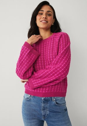 Uśmiechnięta kobieta z długimi ciemnymi włosami, ubrana w teksturowany jasnoróżowy sweter i niebieskie dżinsy, pozująca na tle gładkiej jasnej ściany.