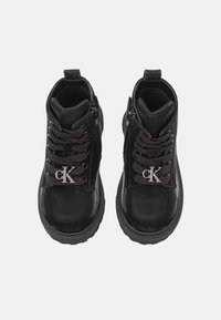 Botines negros con un acabado brillante, puntera redonda, diseño de cordones, cremallera lateral y un acento en forma de logo "CK" plateado en la lengüeta.