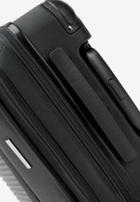 Valise rigide noire avec surface texturée, double fermeture éclair et logo en relief. Elle dispose d'une poignée rétractable et d'une poignée latérale pour un transport facile.