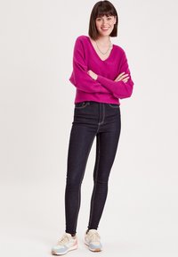 Un pull rose à col en V avec des épaules tombantes, associé à un jean en denim foncé taille haute avec des coutures contrastées, et des baskets de couleur claire.