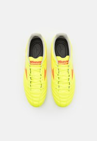 Mizuno nogometne kopačke s svijetlo žutim sintetičkim gornjim dijelom, vezanjem preko čipke, podstavljenim ovratnikom i narančastim detaljima duž strana.