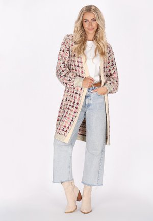 Cardigan lavorato a maglia con motivo geometrico in crema, rosa e viola. Presenta polsini e orlo a coste, apertura frontale, indossato sopra una maglietta bianca e jeans azzurri chiari.