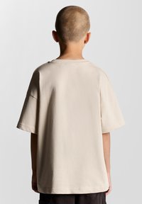 Beige bomulds-T-shirt med en afslappet pasform, korte ærmer og en rund halsudskæring, vist fra bagsiden. Ingen synlige mønstre eller hardware detaljer.