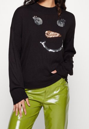 Schwarzer Pullover mit einem Pailletten-Schneemann-Gesicht und glänzenden limettengrünen Hosen, getragen von einer Frau mit der Hand in der Tasche und weiten Ärmeln.